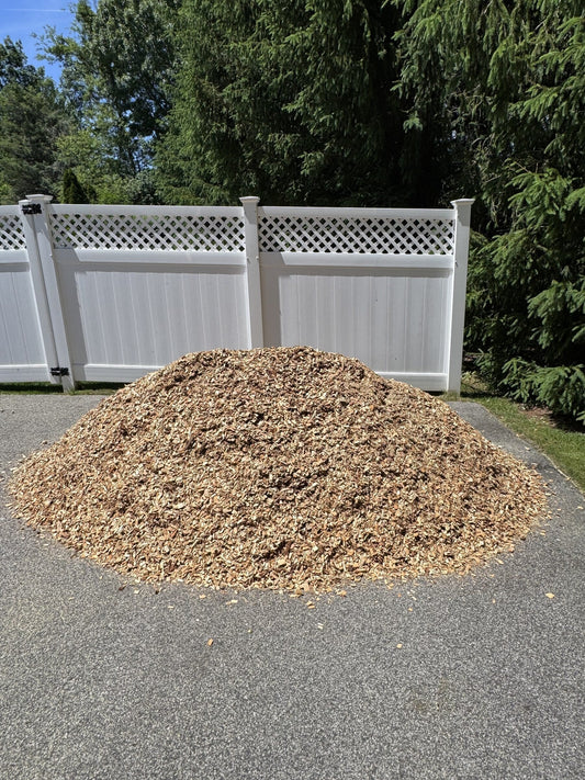 Pile of light tan playground mulch on a residential driveway in front of a white vinyl fence