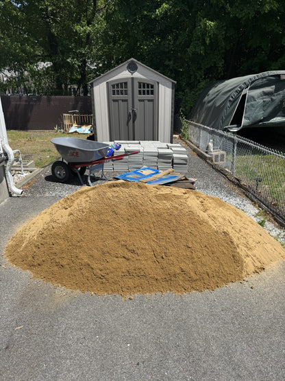 One cubic yard pile of paver sand delivered to a residential driveway in Chelmsford, Massachusetts, staged next to pallets of pavers and a wheelbarrow for a DIY project