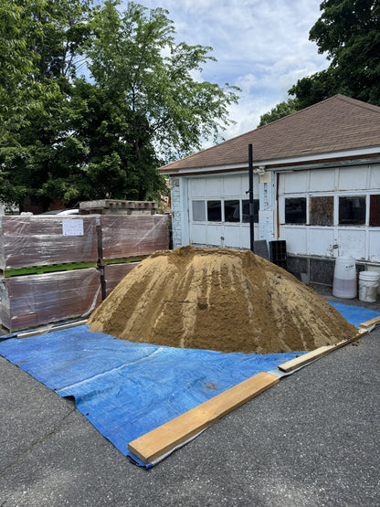 Four cubic yard pile of paver sand delivered to a residential driveway in Lowell, Massachusetts, staged next to pallets of pavers for a backyard patio install